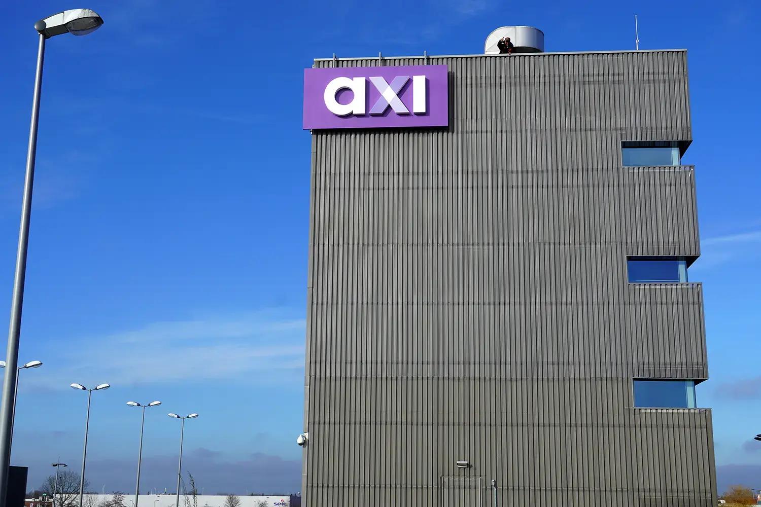 Grote paarse gevelreclame met witte doosletters "axi" hoog gemonteerd op de grijze gevel van een modern bedrijfspand langs een bedrijventerrein.