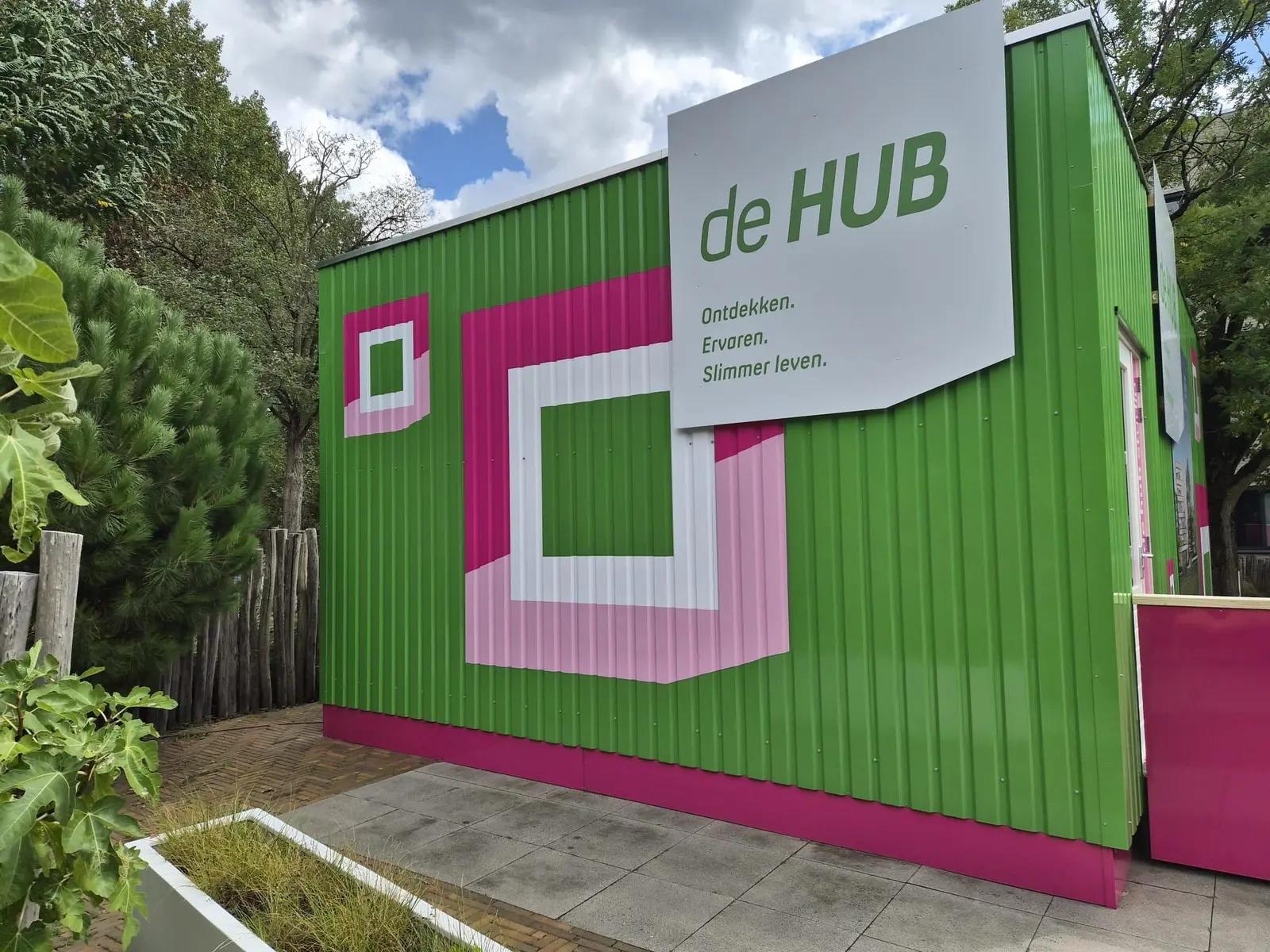 Zijwand van de vaste HUB met witte en roze vormen en de boodschap Ontdekken, Ervaren, Slimmer leven.