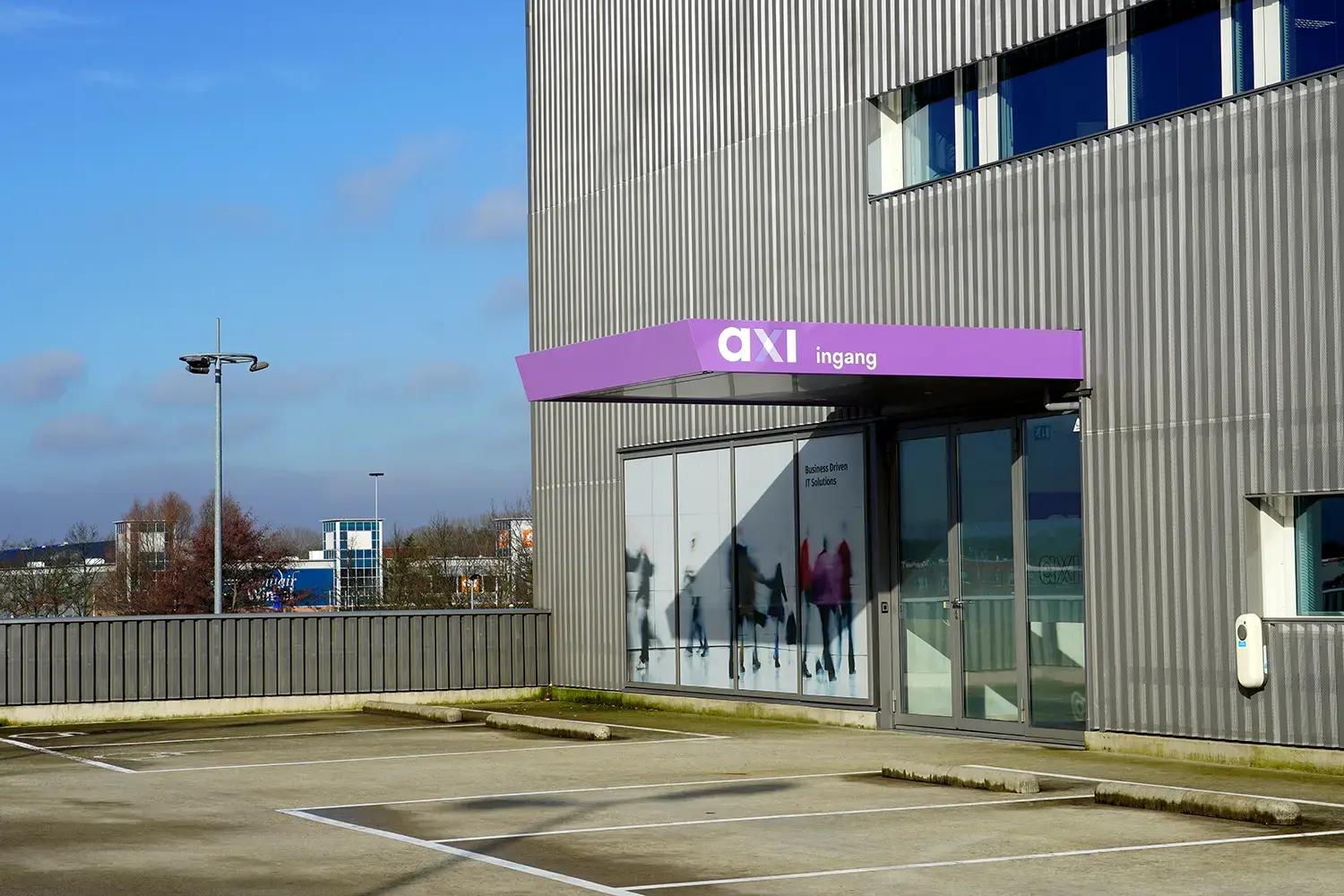 Paarse luifelreclame met witte belettering "axi ingang" boven de entree van een modern bedrijfspand met grijze gevelbekleding en parkeerplaats.