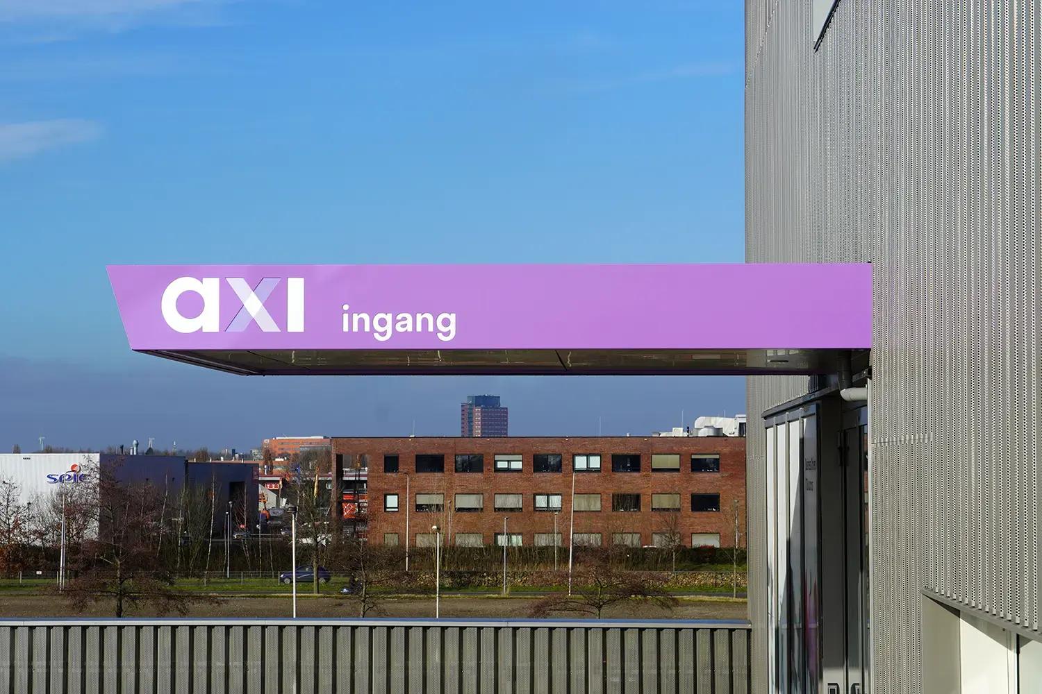 Paarse luifelreclame met witte belettering "axi ingang" aan de gevel van een modern bedrijfspand, zichtbaar boven de entree met uitzicht op een bedrijventerrein.
