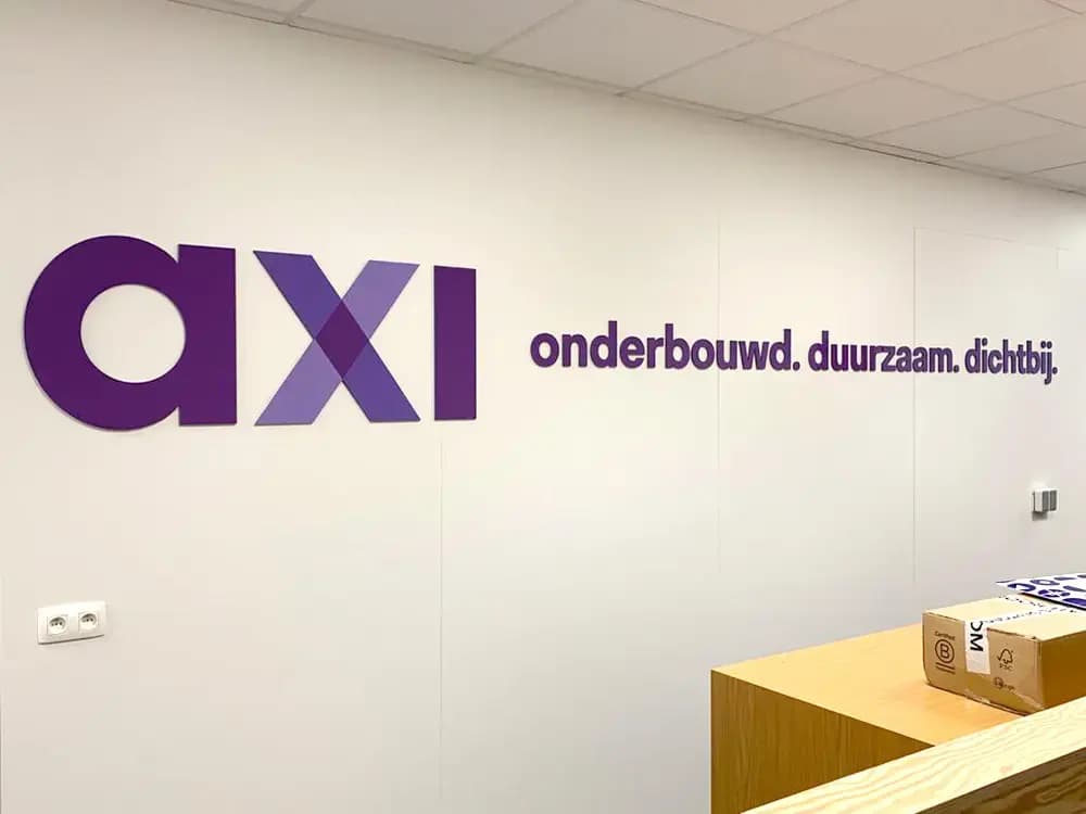 Witte kantoorwand met paarse interieurbelettering van het axi-logo en de slogan "onderbouwd. duurzaam. dichtbij." achter een houten balie.