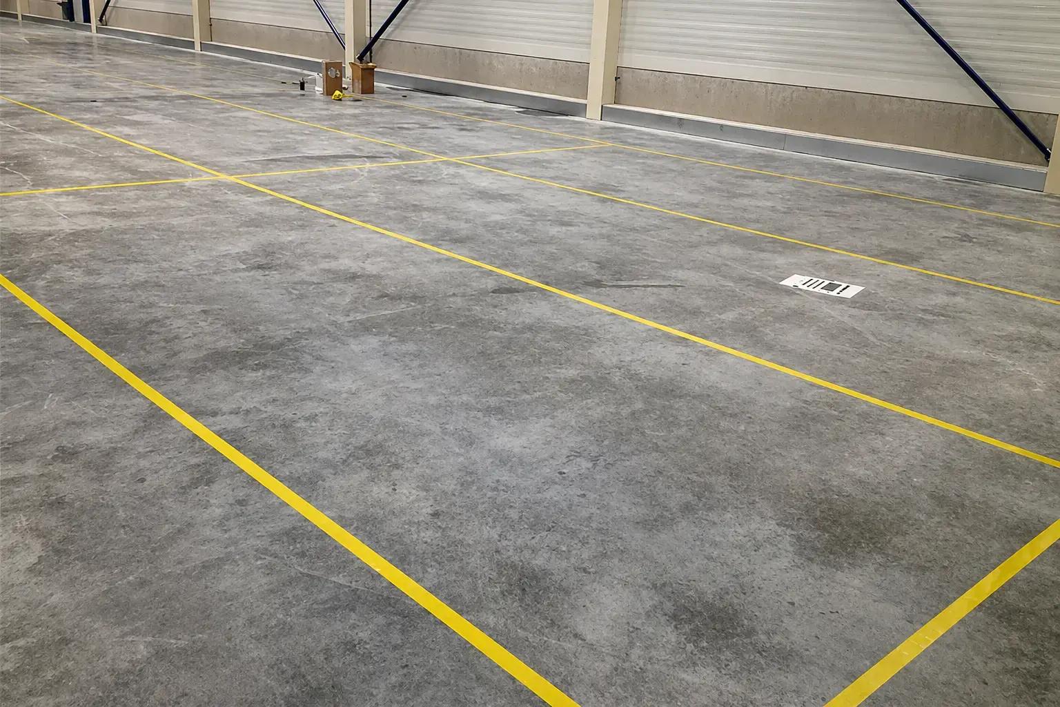 Gele vloerbelijning op een betonnen magazijnvloer met rechthoekige vakindeling, toegepast als duidelijke veiligheidsmarkering en logistieke routeaanduiding.