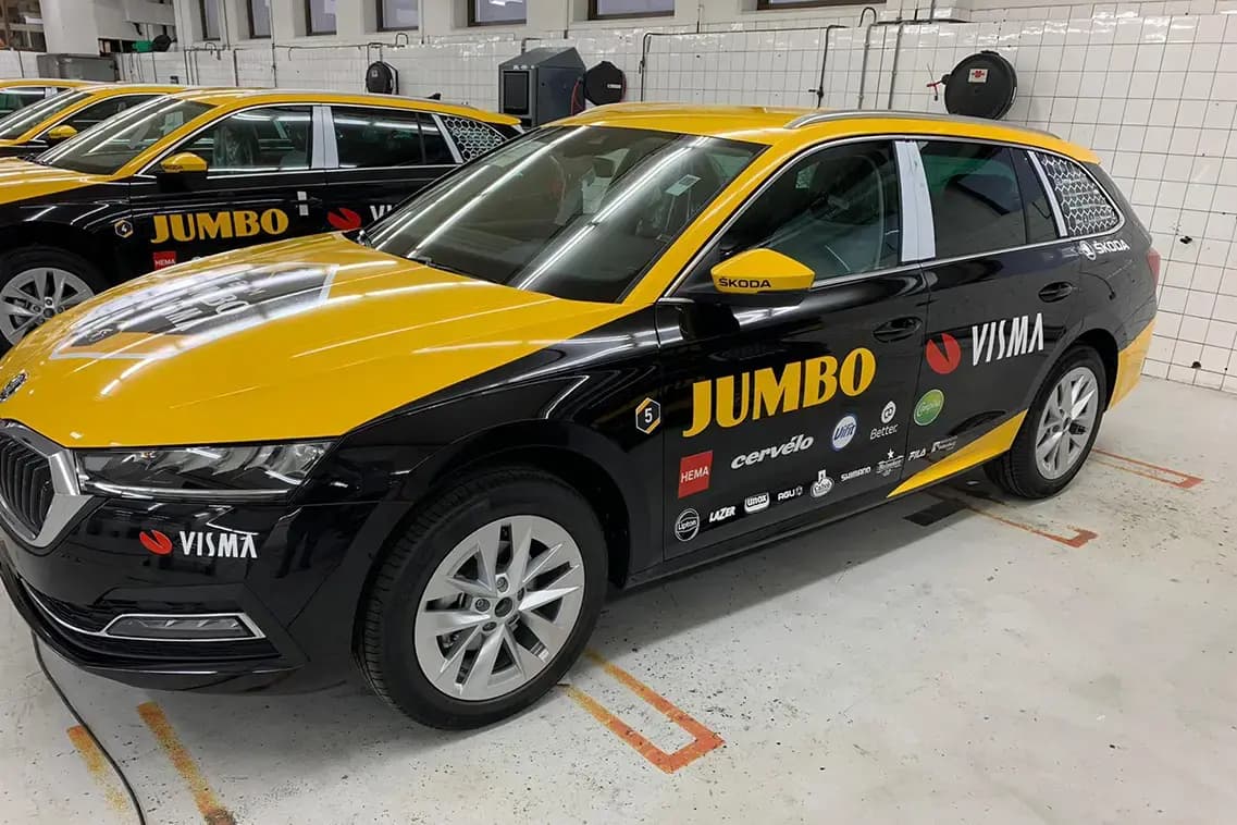 Zwarte en gele Škoda stationwagen met full-colour voertuigbelettering voor Jumbo Visma, voorzien van sponsorlogo’s op de zijkant en gefotografeerd in een werkplaats.