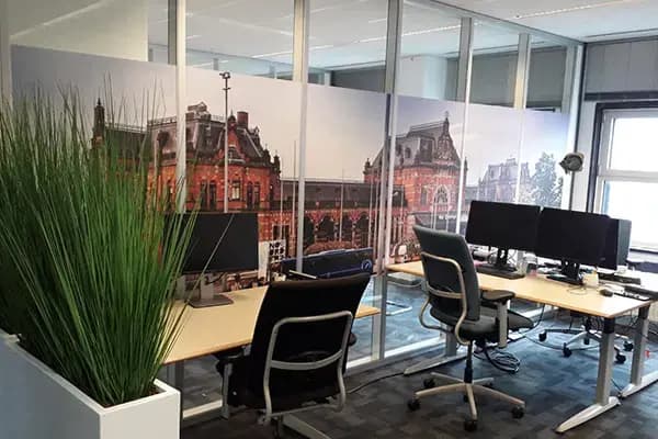 Kantoorruimte met glazen scheidingswand voorzien van full-colour glasfolie met stadsgezicht, toegepast als privacyfolie tussen werkplekken met bureaus en computerschermen.
