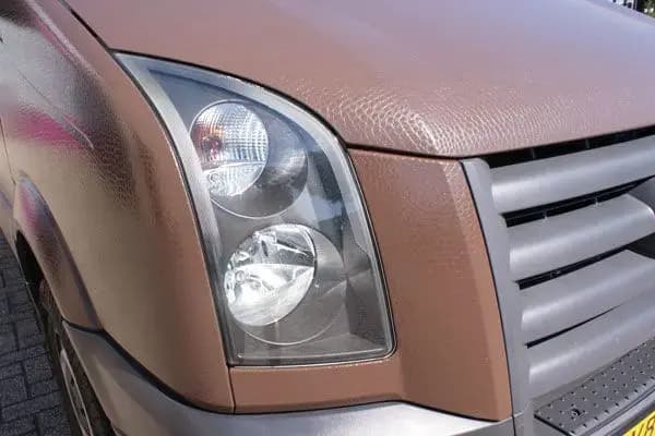 Close-up van de voorzijde van een bestelbus met bruine carwrap, zichtbare folietextuur, koplampdetail en zilverkleurige grille.