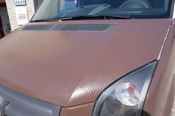 Close-up van de motorkap van een bestelbus met bruine carwrap, zichtbare folietextuur, ventilatierooster en koplampdetail aan de voorzijde.