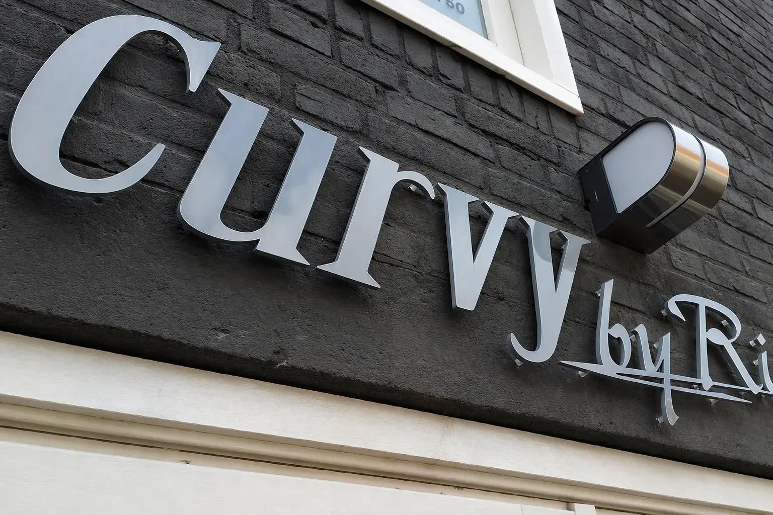 Zilverkleurige 3D gevelletters met merkbelettering op een donker gemetselde gevel, afgewerkt met een wandlamp boven de entree.