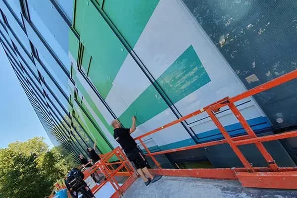 Montage van full-colour raamfolie op een glazen gevel met grote grafische vlakken, uitgevoerd vanaf oranje hoogwerkers aan een modern kantoorgebouw.