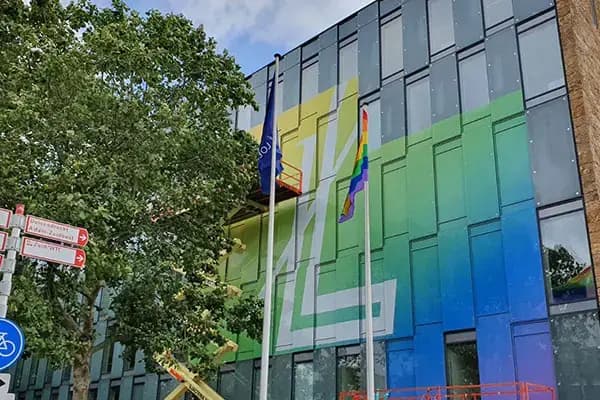 Glazen gevel van een modern kantoorgebouw met full-colour raamfolie in groen, blauw en geel, gecombineerd met grote witte vlakletters en vlaggen voor het pand.