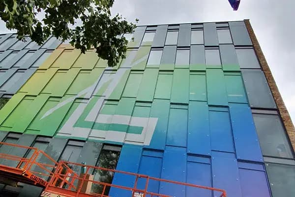 Glazen gevel van een modern kantoorgebouw met full-colour raamfolie in groen- en blauwtinten, voorzien van grote witte grafische vormen en gefotografeerd tijdens montage met een hoogwerker.