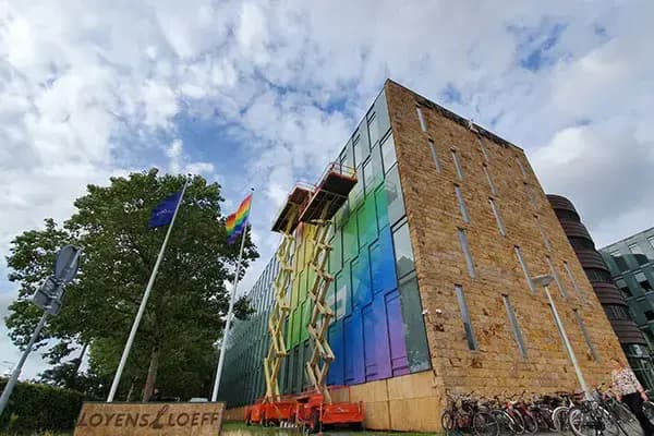 Modern kantoorgebouw met glazen hoekgevel voorzien van regenboogkleurige raamfolie, gefotografeerd tijdens montage met hoogwerkers en vlaggen voor het pand.