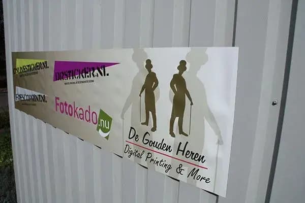 Een grote witte magneetsticker op een grijze metalen wand met daarop verschillende logo's van printbedrijven, waaronder "De Gouden Heren Digital Printing & More".