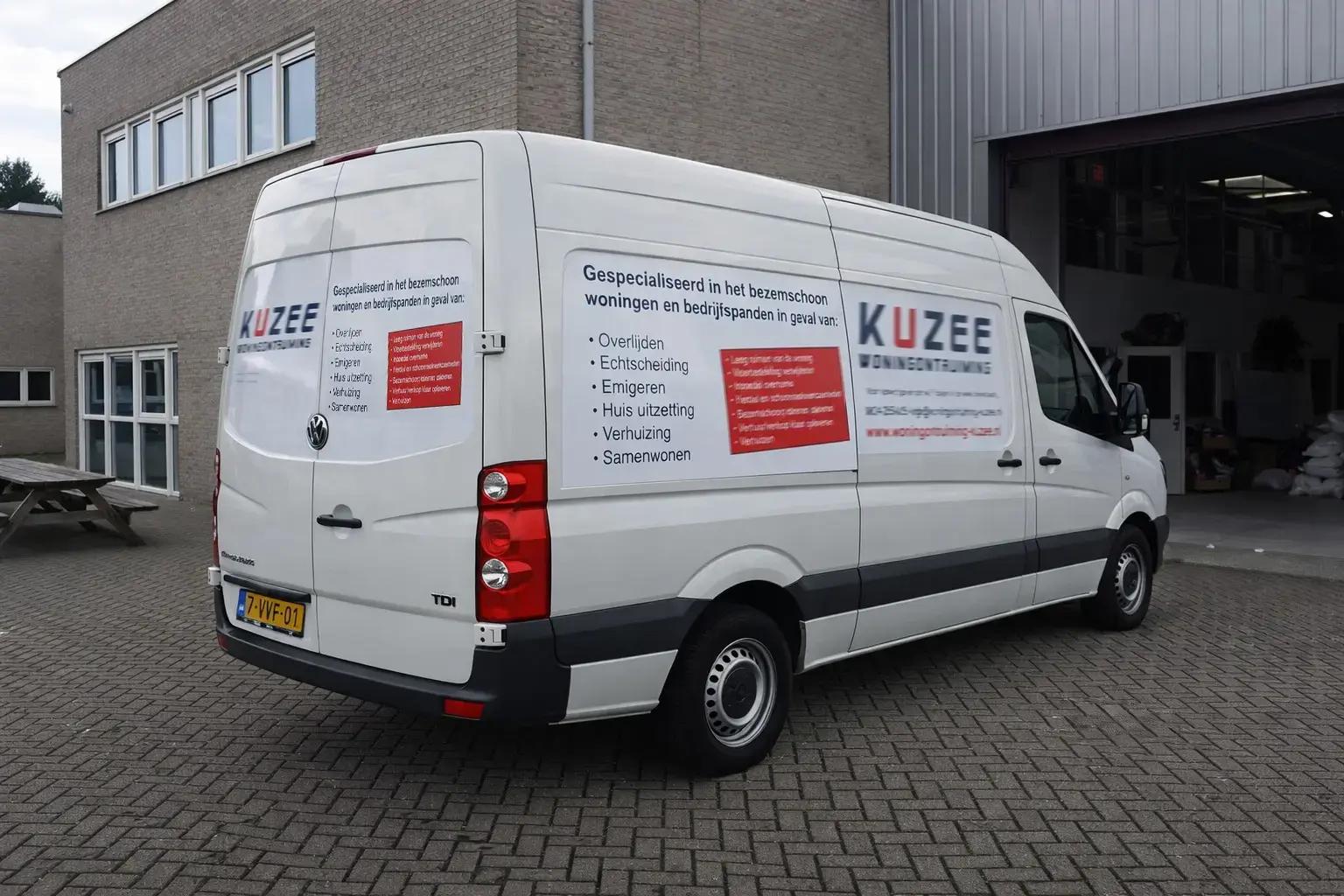 Witte bestelbus met full-colour voertuigbelettering van KUZEE Woningontruiming, voorzien van logo, dienstenoverzicht en rode tekstvlakken, geparkeerd bij een bedrijfspand.