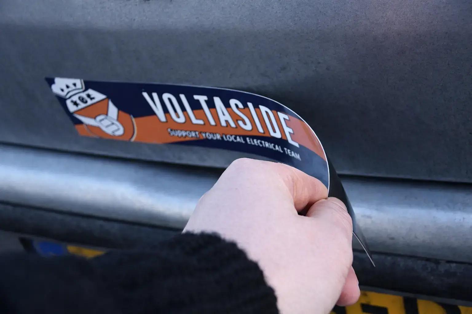 Hand brengt een gebogen voertuigsticker met het logo en de tekst ‘VOLTASIDE’ aan op de grijze metalen achterkant van een voertuig.
