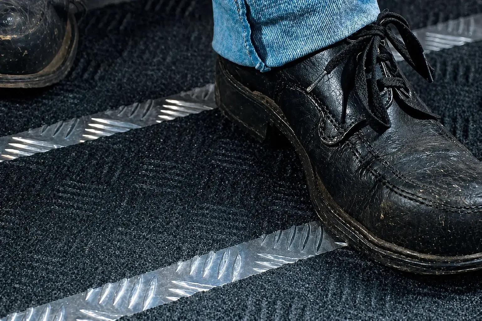 Close-up van zwarte schoenen op een donkere vloermat met zilverkleurige antislipstrips, toegepast als veiligheidsmarkering en slippreventie op een looproute.