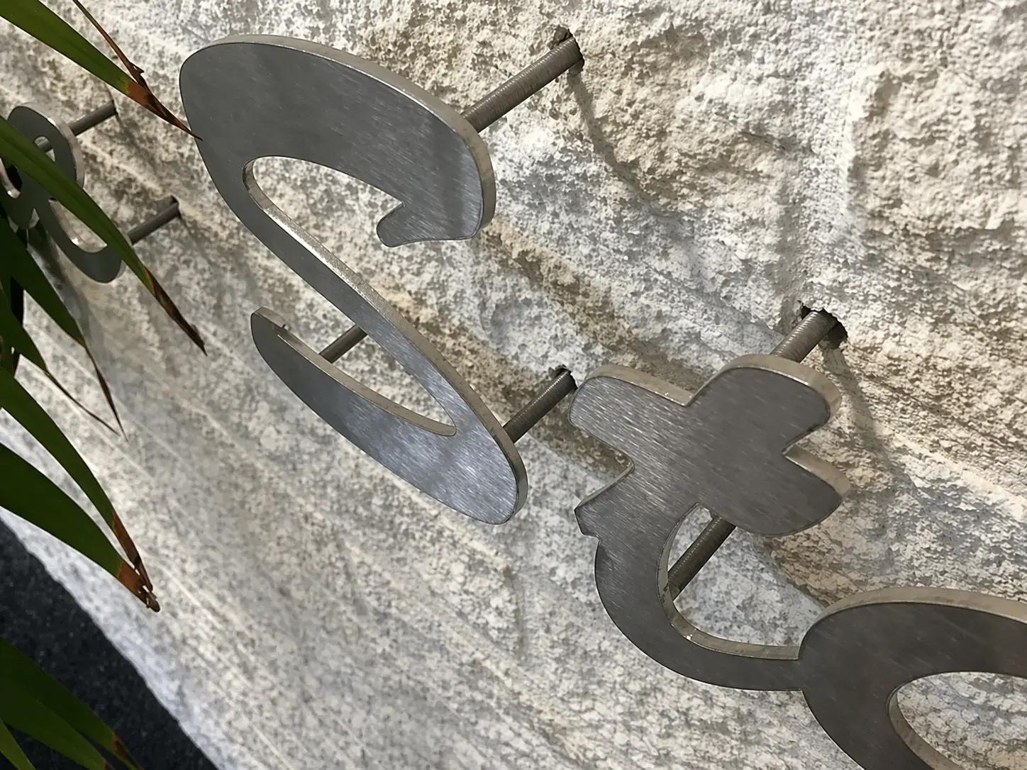 Close-up van geborstelde metalen gevelletters op afstandhouders, gemonteerd op een ruwe natuurstenen wand als luxe 3D-belettering.