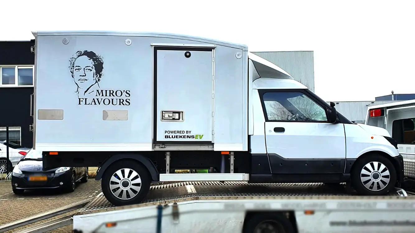 Witte bakwagen met voertuigbelettering voor Miro’s Flavours, voorzien van zwarte merknaam, portretillustratie en sponsorvermelding op de zijkant van de laadbak.