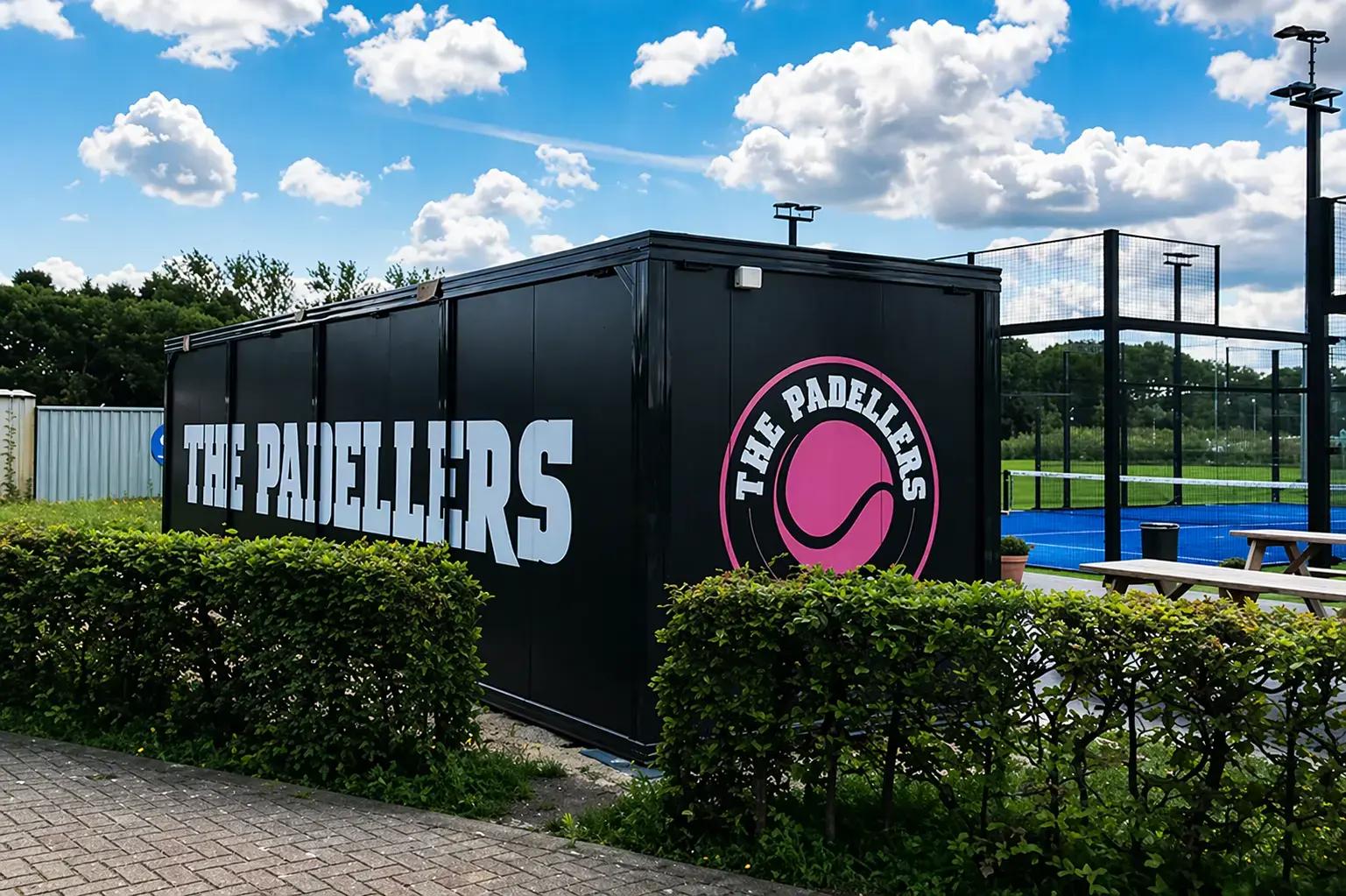 Zwarte containerunit met grote The Padellers-belettering en rond logo, geplaatst naast blauwe padelbanen als opvallende buitenreclame.
