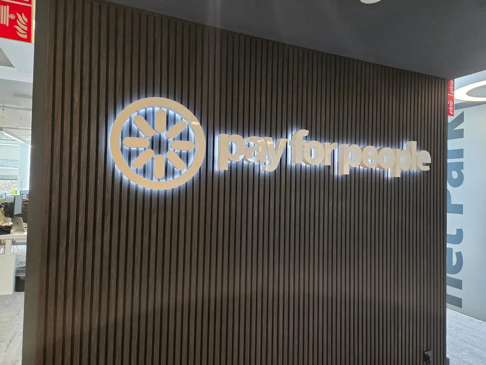 White halo-lit PayforPeople lettering on a dark slatted wall in the office.