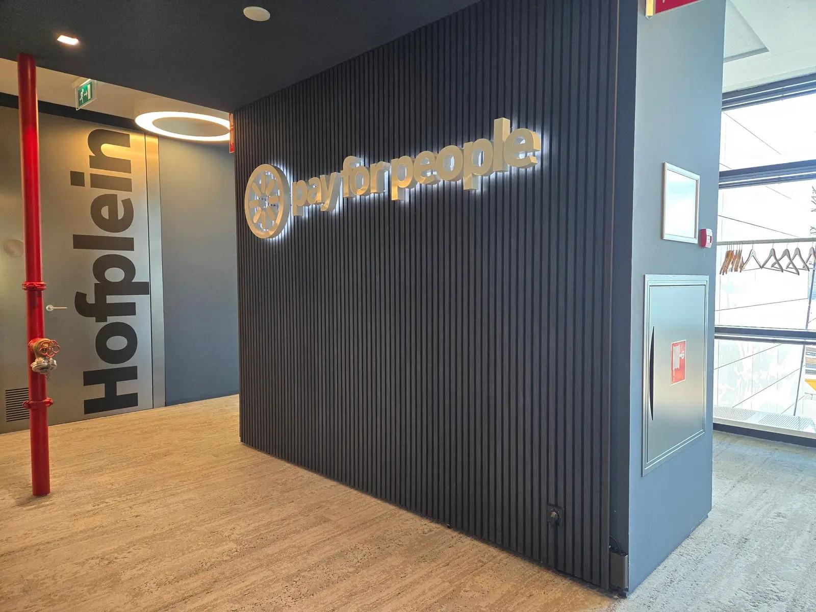 Verlicht PayforPeople-logo op de lattenwand naast de doorgang met de ruimtenaam Hofplein.