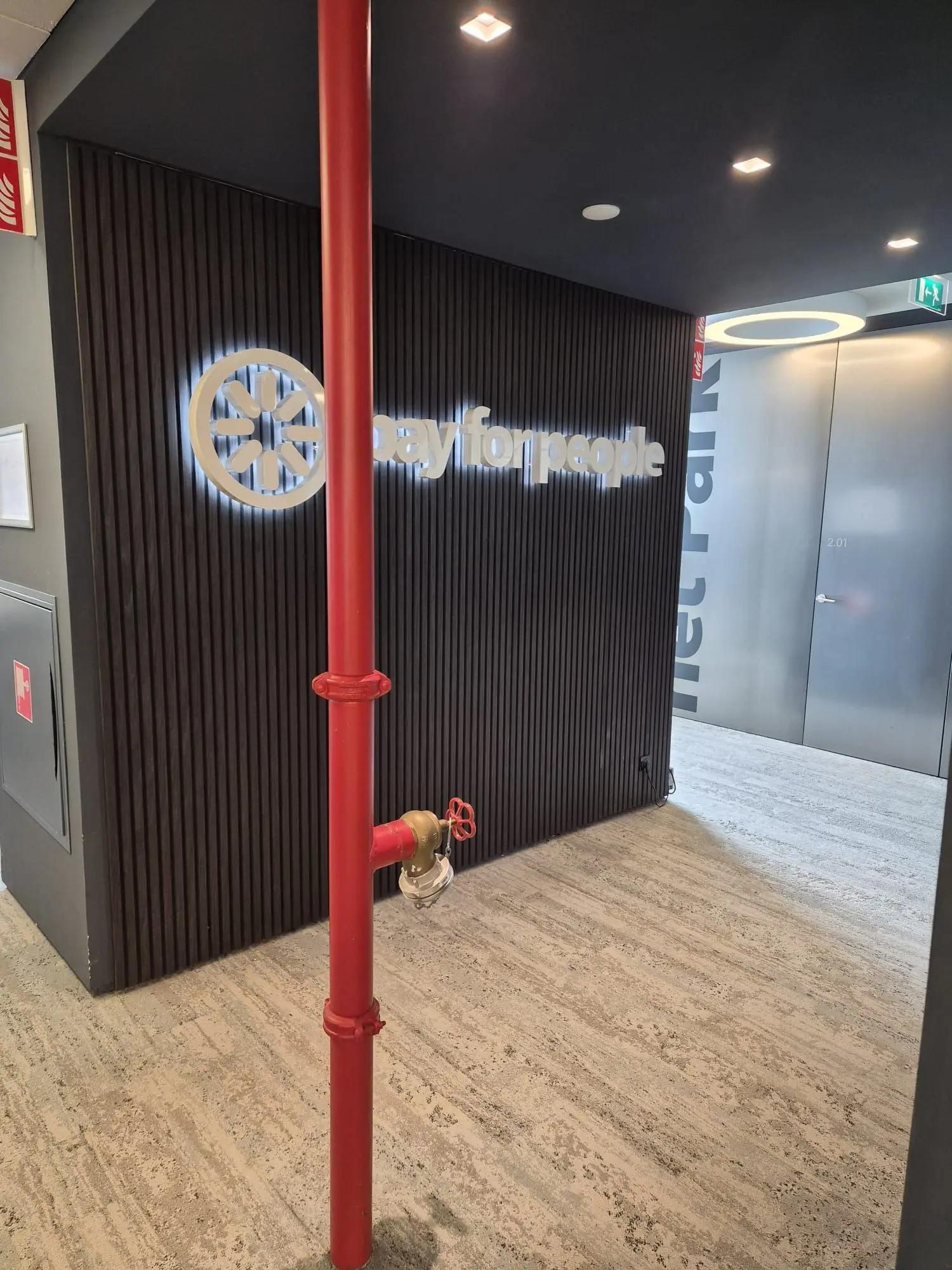 PayforPeople-lichtreclame van opzij gezien, met reliëfletters en gloed tegen de lattenwand.