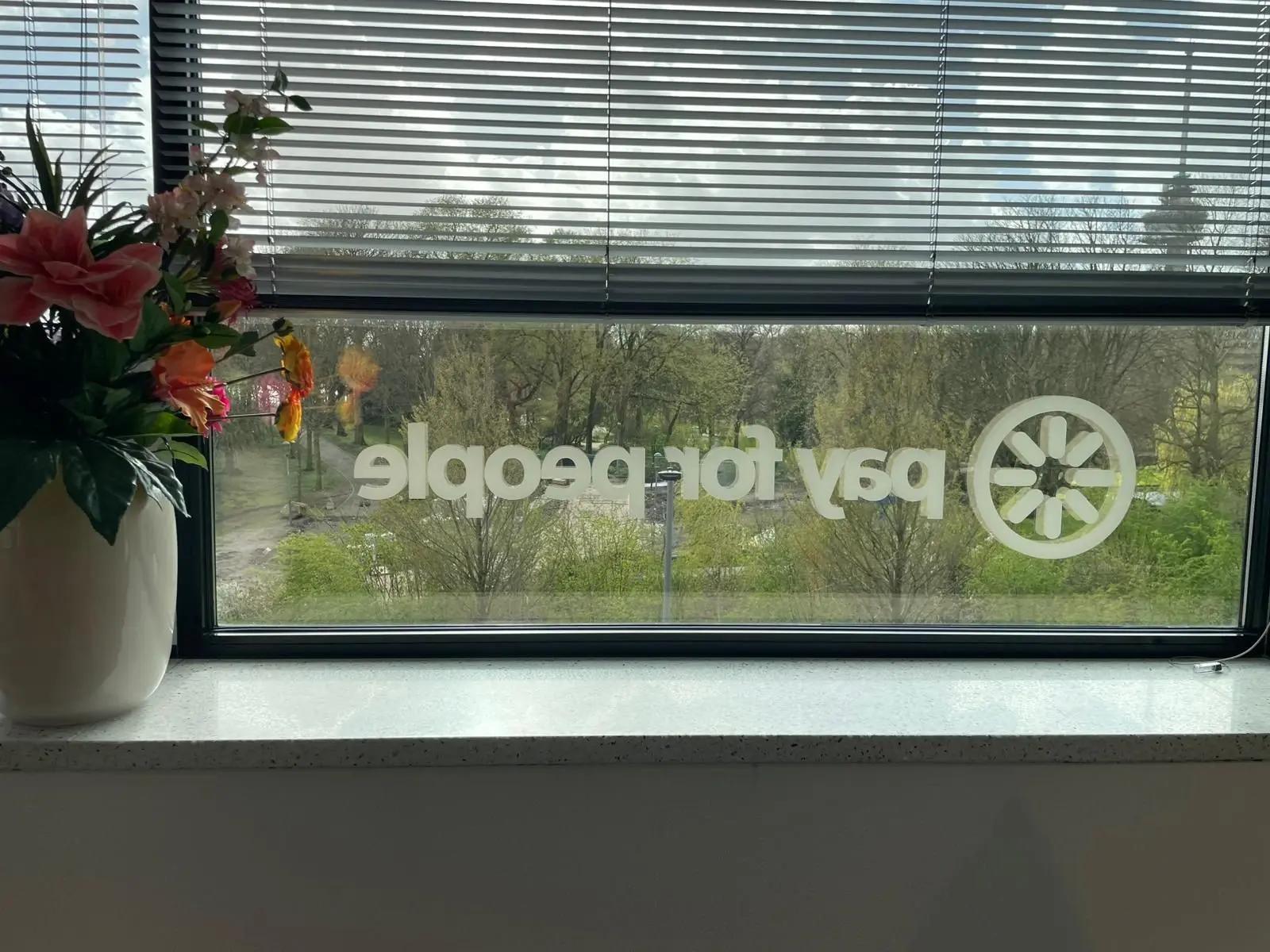 PayforPeople-logo op het raam naast een bloemstuk, met zicht op bomen buiten.
