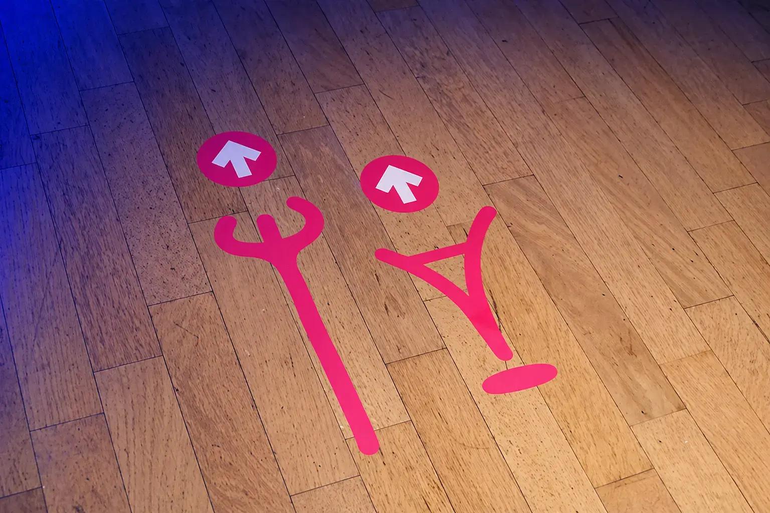Roze vloerbelettering met pijlstickers op een houten vloer als wayfinding-signing voor looprichting en afstandsaanduiding.