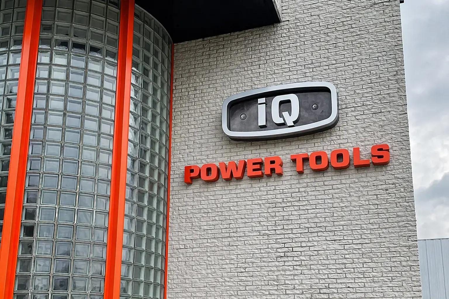 Gevelreclame op een lichtgrijze bakstenen gevel met iQ-logo in een omlijst paneel en rode 3D doosletters met de tekst POWER TOOLS naast een glazen hoekgevel.