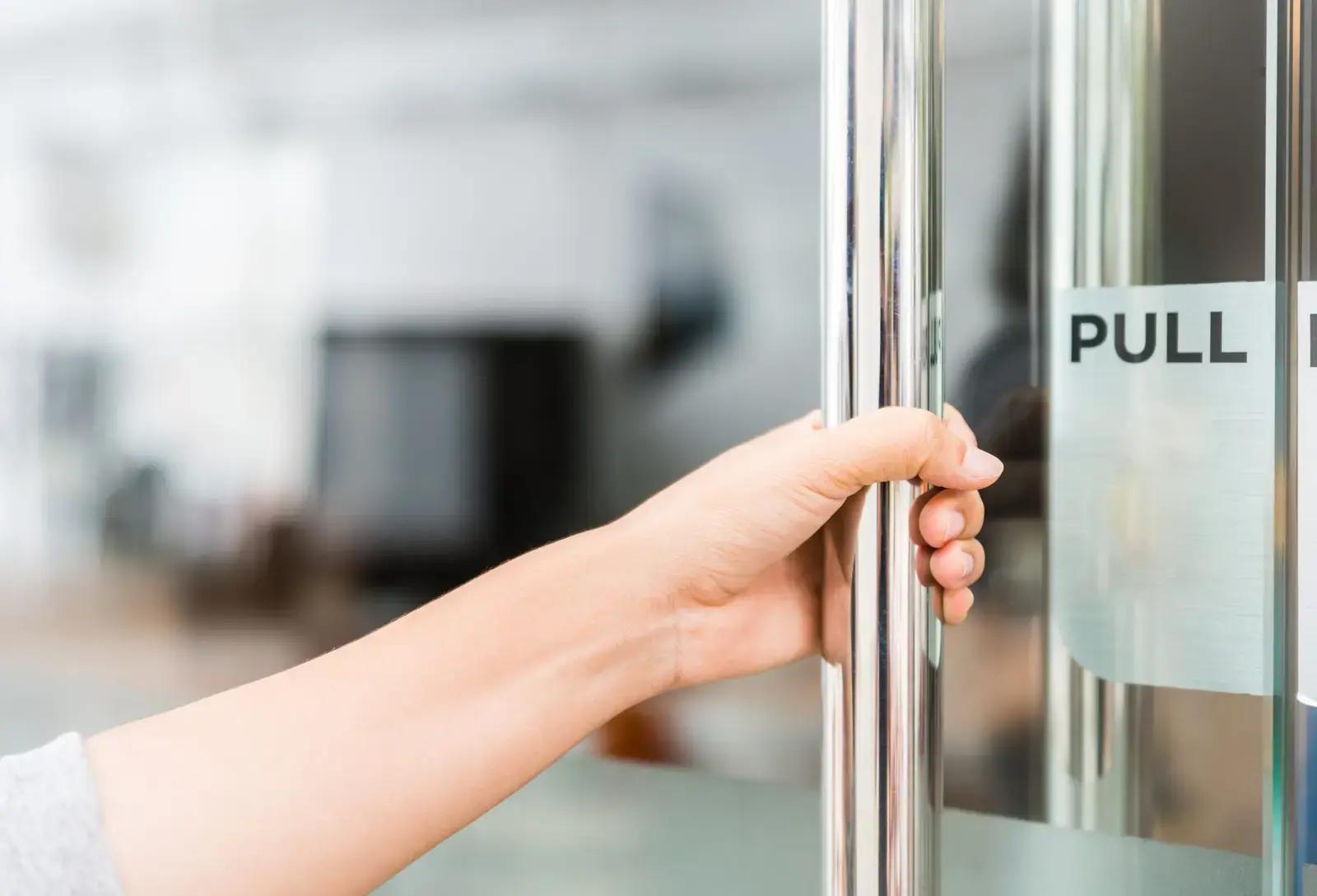 Hand aan een metalen glazen deurklink met duidelijke PULL-deursticker, toegepast als functionele bewegwijzering op een glazen kantoorentree.