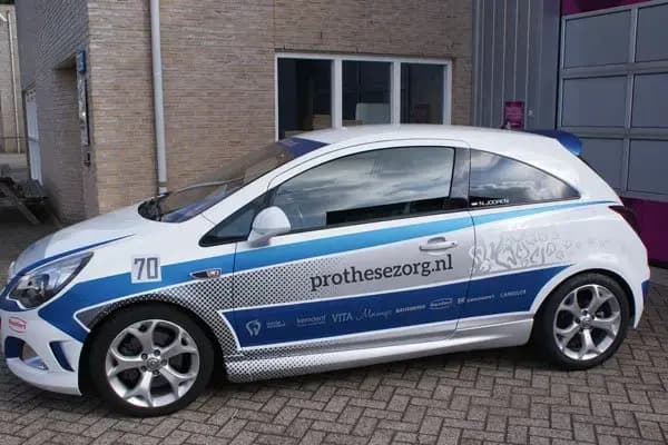 Zijaanzicht van een witte personenauto met blauwe en grijze voertuigbelettering, prothesezorg.nl op het portier en sponsorlogo’s langs de onderzijde, geparkeerd op een bedrijventerrein.