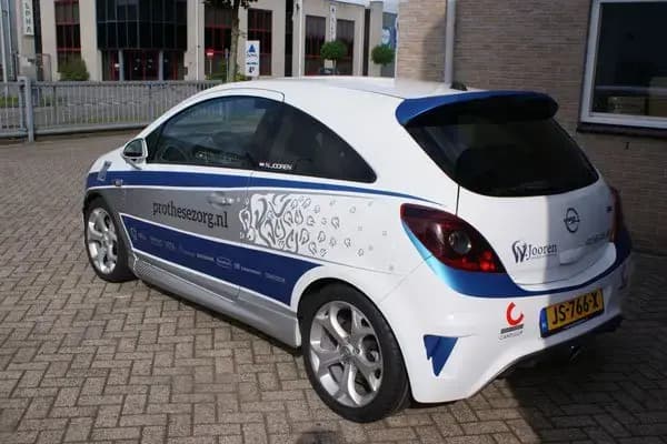 Achterzijde van een witte personenauto met blauwe en grijze voertuigbelettering, websitevermelding en decoratieve bestickering op de zijpanelen, geparkeerd op een bedrijventerrein.