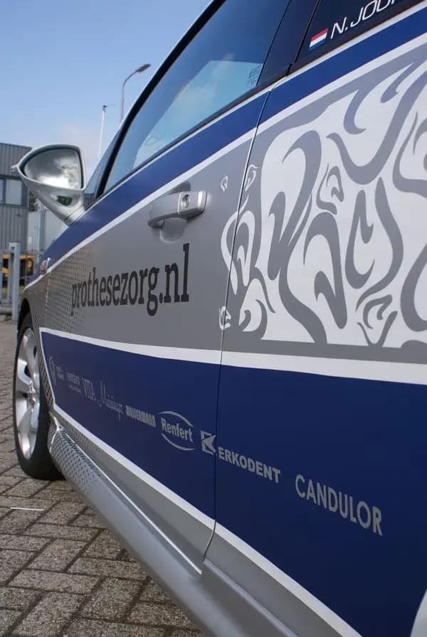 Close-up van blauwe en grijze voertuigbelettering op een witte personenauto, met prothesezorg.nl, decoratief patroon en sponsorlogo’s op het portier.