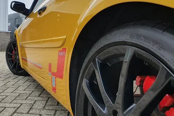 Close-up van een gele personenauto met sportieve voertuigbelettering op de dorpel, zwarte lichtmetalen velg en rode remklauw.