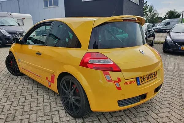 Gele driedeurs hatchback met sportieve voertuigbelettering, rode striping op de zijkant en zwarte lichtmetalen velgen, geparkeerd op een bedrijventerrein.
