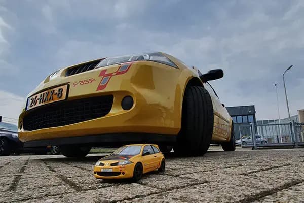 Gele personenauto met sportieve voertuigbelettering en rode striping op de voorzijde, gefotografeerd vanaf laag standpunt met een bijpassend miniatuurmodel op de bestrating.