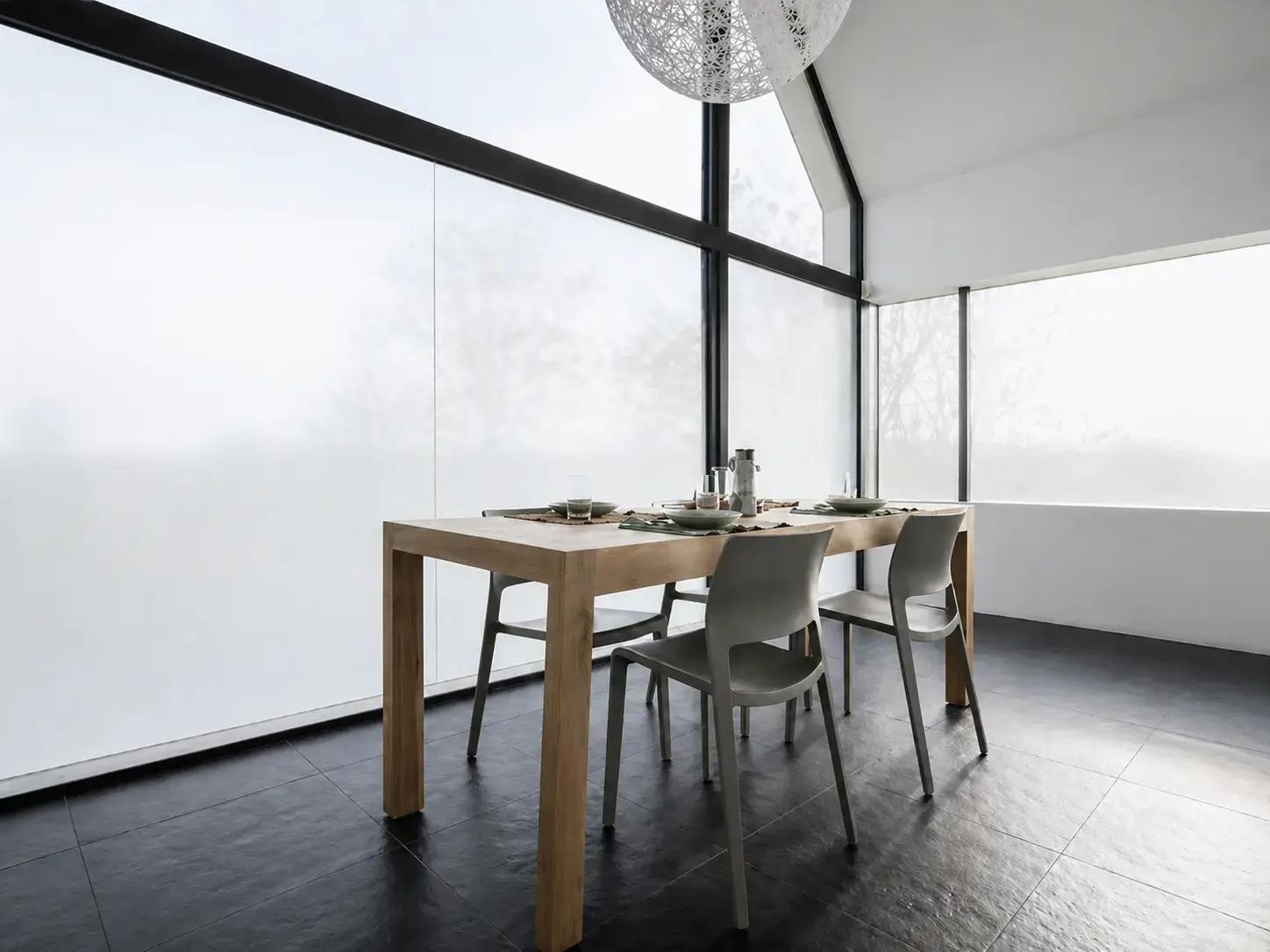 Matte privacyfolie op grote raampartijen in een moderne eetruimte met houten tafel en zwarte kozijnen.