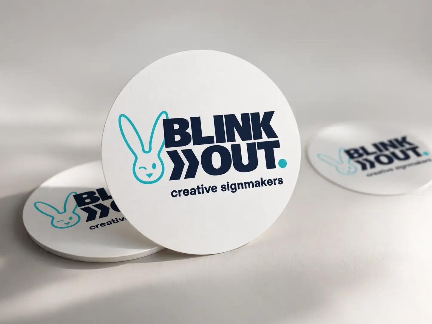 Witte ronde onderzetters met BLINK OUT creative signmakers-logo, gepresenteerd als promotioneel relatiegeschenk op een neutrale achtergrond.