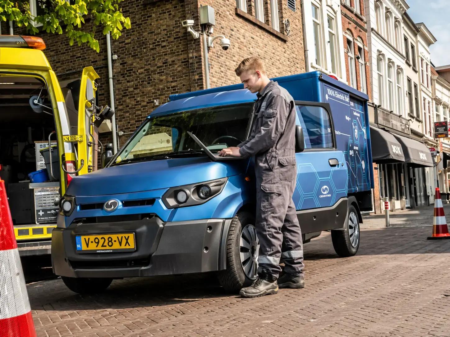 Blauwe bedrijfswagen met full-colour voertuigbelettering en bestickering geparkeerd in een winkelstraat, terwijl een monteur met laptop naast het voertuig staat.
