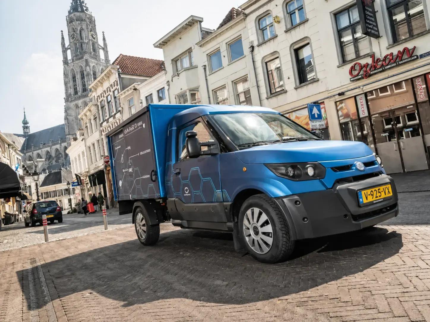 Blauwe bakwagen met voertuigbelettering en grafische bestickering in een historisch stadscentrum, met winkels en kerk op de achtergrond.