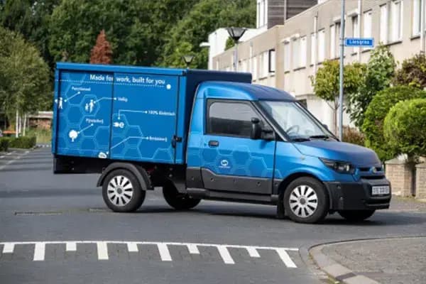Blauwe bakwagen met voertuigbelettering en informatieve zijpaneelbestickering rijdt door een woonwijk, met hexagonpatroon en tekst op de laadbak.