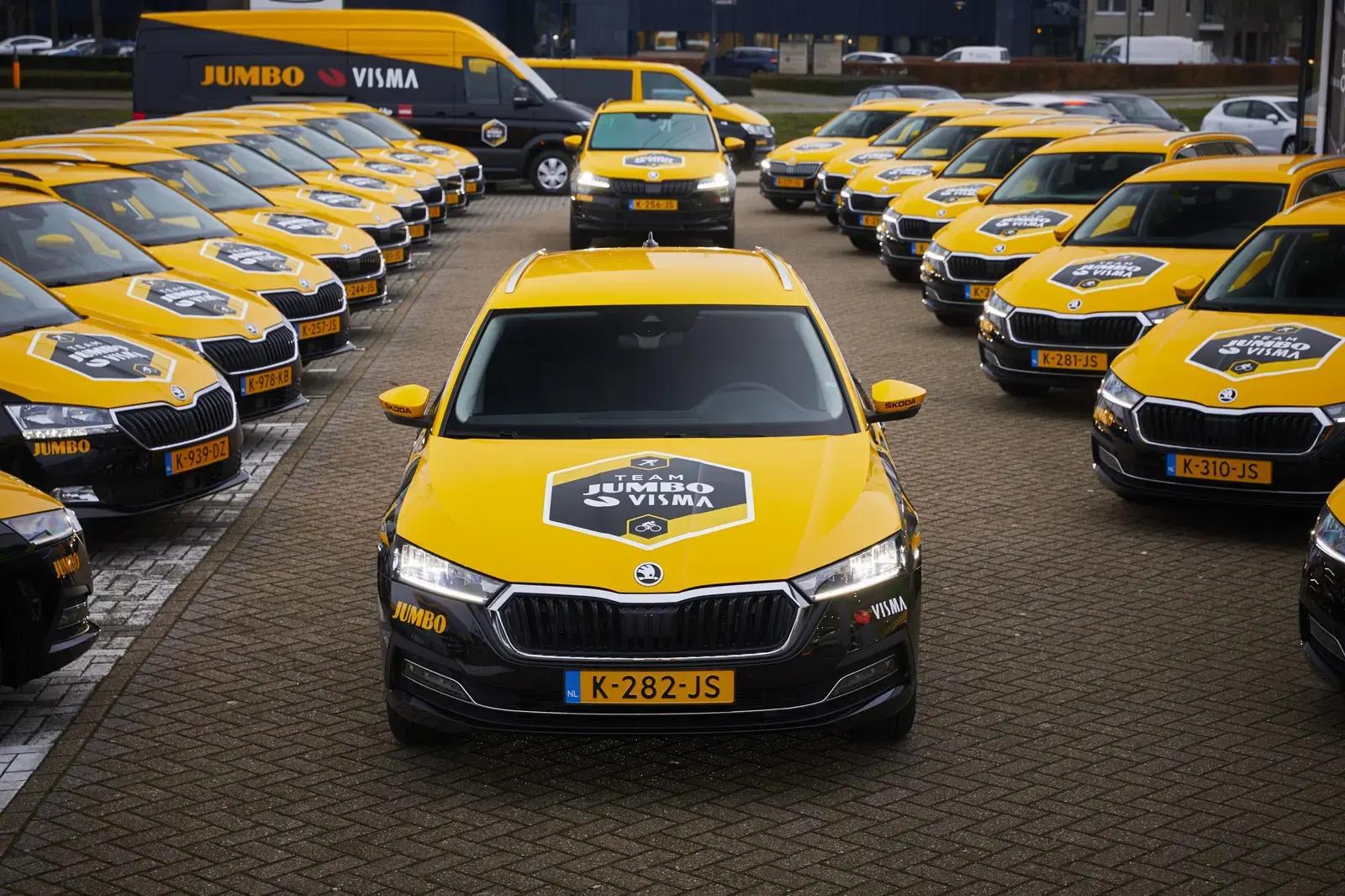 Grote vloot gele en zwarte Škoda-teamauto’s en een bestickerde Jumbo Visma-bus met full-colour voertuigbelettering, opgesteld op een parkeerterrein.