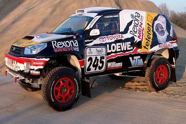 Zwarte, witte en rode off-road rallyauto met volledige voertuigwrap, sponsorbelettering en grote logo’s op de zijkanten, gefotografeerd op zandterrein.