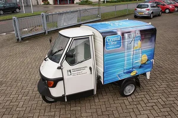 Compacte bezorgscooter met gesloten laadbak en full-color voertuigbelettering voor Crystal Clear, voorzien van productafbeeldingen op de zijkant en geparkeerd op een bedrijventerrein.