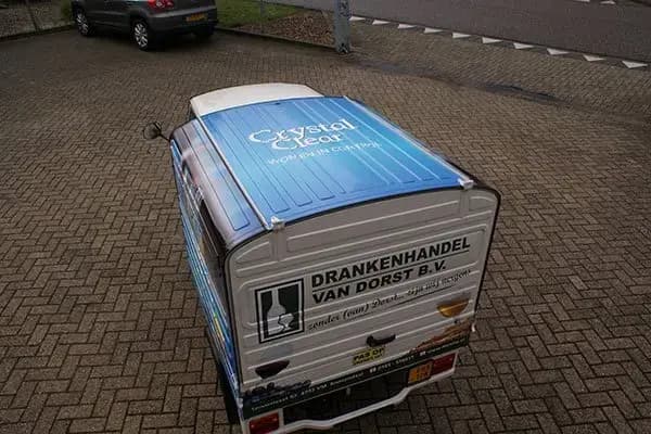 Bovenaanzicht van een compacte bezorgscooter met gesloten laadbak, voorzien van blauwe dakbestickering voor Crystal Clear en achterbelettering van Drankenhandel Van Dorst B.V. op een bedrijventerrein.