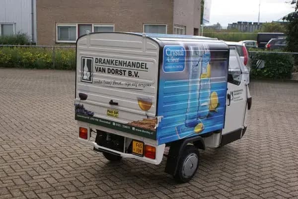 Achterzijde van een compacte bezorgscooter met gesloten laadbak, voorzien van voertuigbelettering voor Drankenhandel Van Dorst B.V. en full-color reclameprint van Crystal Clear op de zijpanelen.