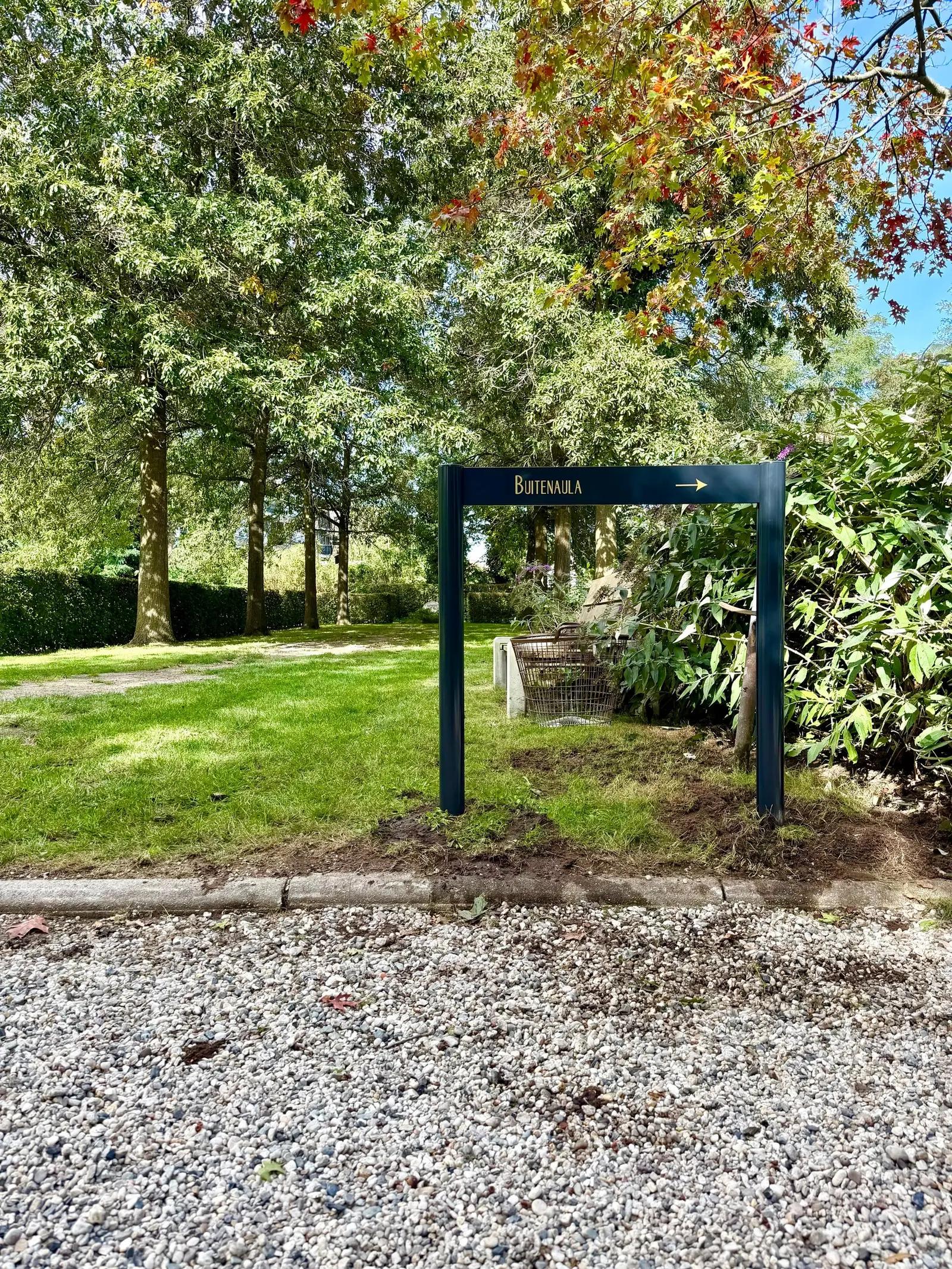 Dark entrance sign with gold-coloured lettering beside a path in a green setting.