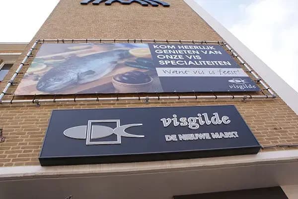 Gevel van een viswinkel met groot Visgilde-gevelbord en gespannen full-color bannerframe met promotionele visafbeelding en actietekst op de bakstenen voorgevel.