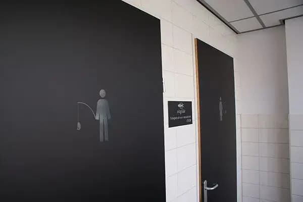 Zwarte toiletdeuren met subtiele sanitaire pictogrammen en wayfinding-bewegwijzering op een wit betegelde wand in een bedrijfsinterieur.