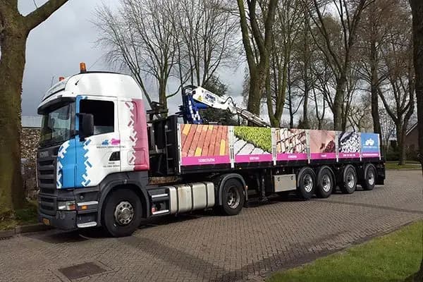 Witte vrachtwagen met cabinebelettering in blauw en roze, full-colour trailerreclame met productfoto’s en tekstpanelen, geparkeerd langs een straat met bomen.
