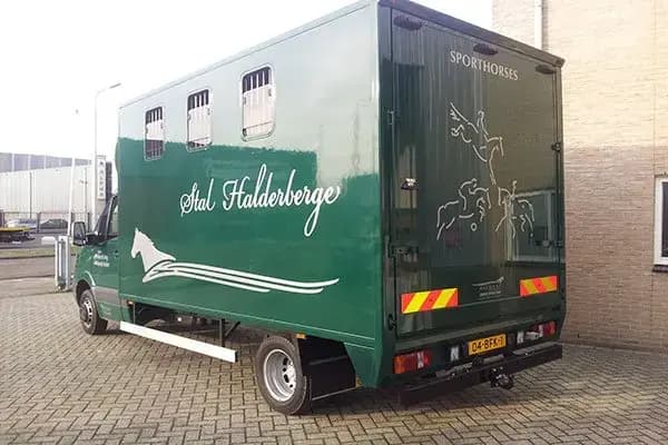 Groene paardenvrachtwagen met Stal Halderberge-belettering, witte paardenafbeelding op de zijkant en Sporthorses-tekst op de achterdeuren, geparkeerd op een bedrijventerrein.